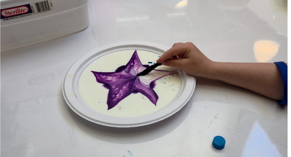 A hand drawing a purple star through dye floating on milk